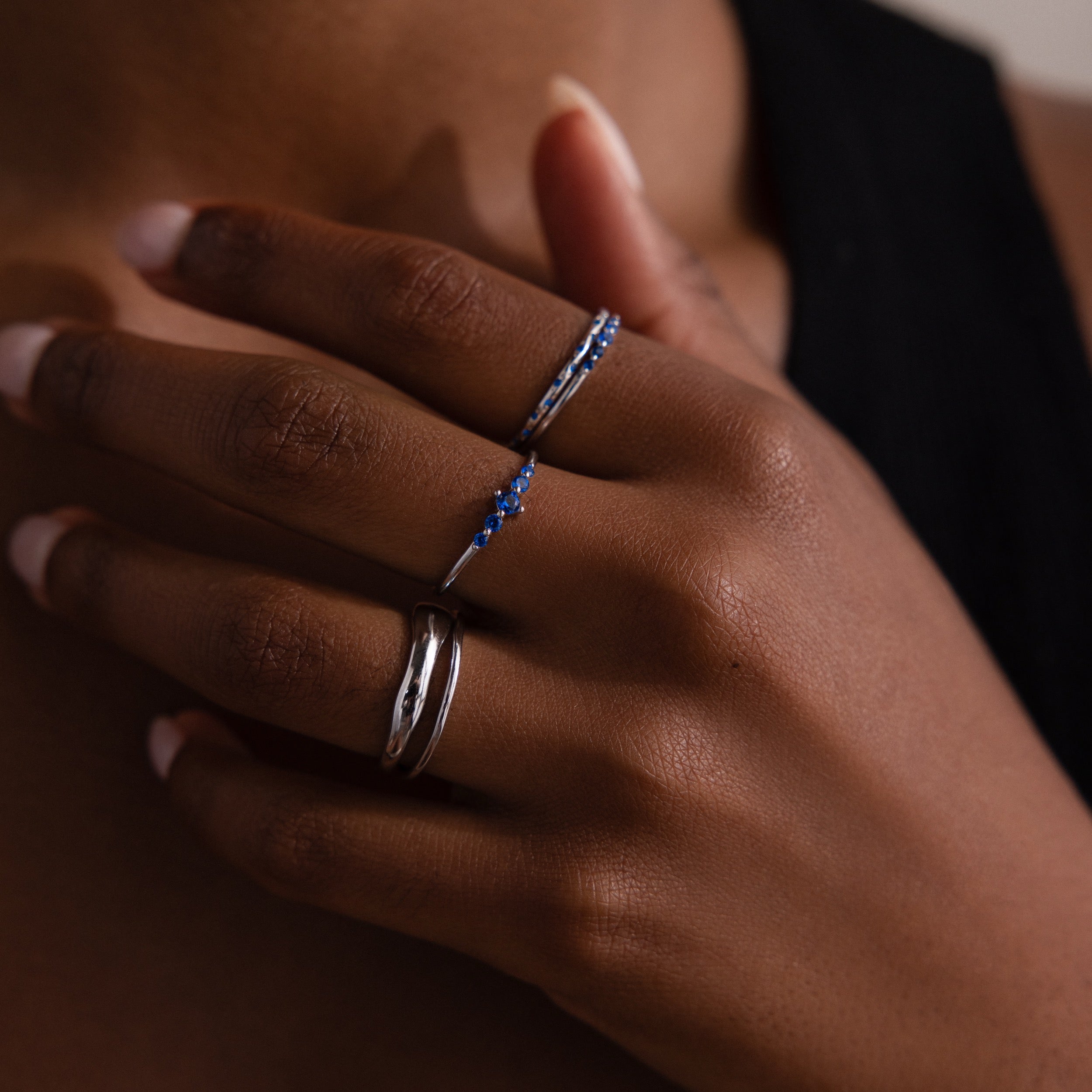 Close-up of a model wearing three rings with blue stones and a set of 2 other duo form silver rings on a dark background