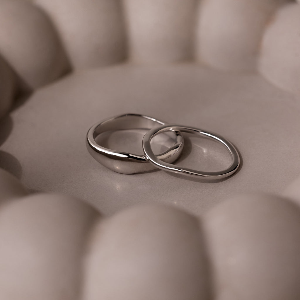 The Duo Form Ring Set in Silver features two sculptural rings resting together on a soft, round beige surface with gentle, curved backgrounds.