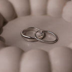 The Duo Form Ring Set in Silver features two sculptural rings resting together on a soft, round beige surface with gentle, curved backgrounds.