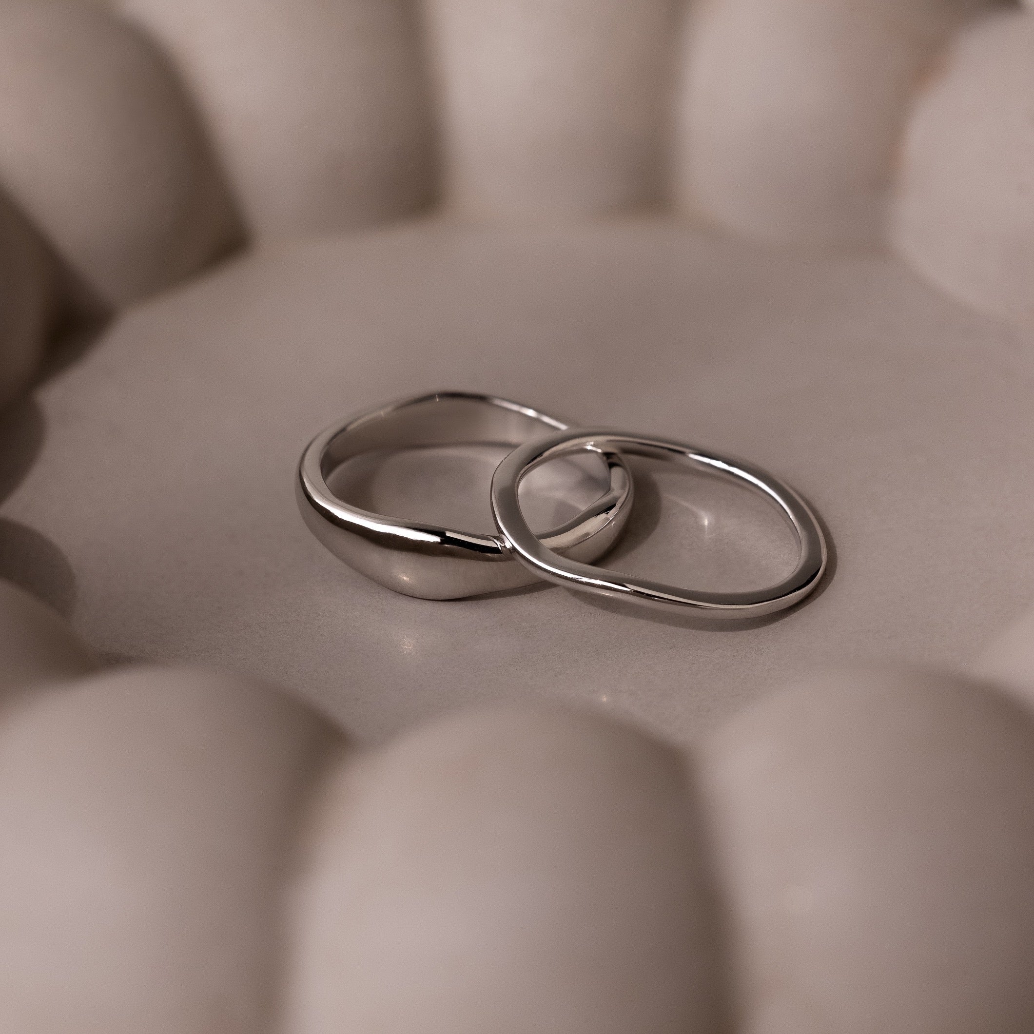 The Duo Form Ring Set in Silver features two sculptural rings resting together on a soft, round beige surface with gentle, curved backgrounds.