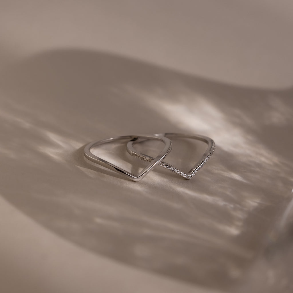 The Curve Stacking Set in Silver features two delicate rings with angular designs, ideal for a modern stack and shown casting soft shadows on a neutral background.