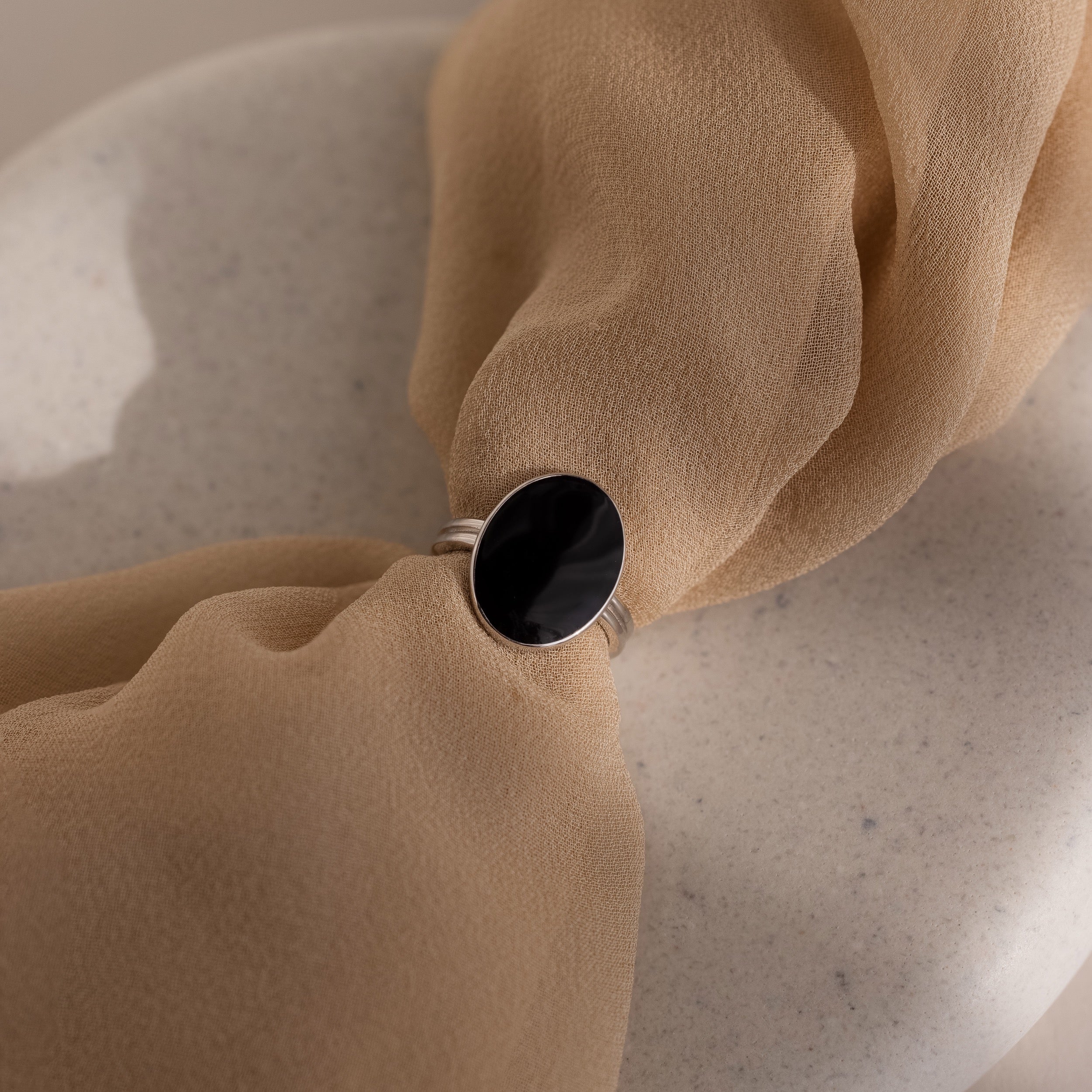 The Midnight Hour Onyx Ring in Silver features a large black oval stone and adjustable band, shown on soft beige fabric for a vintage-glam look.