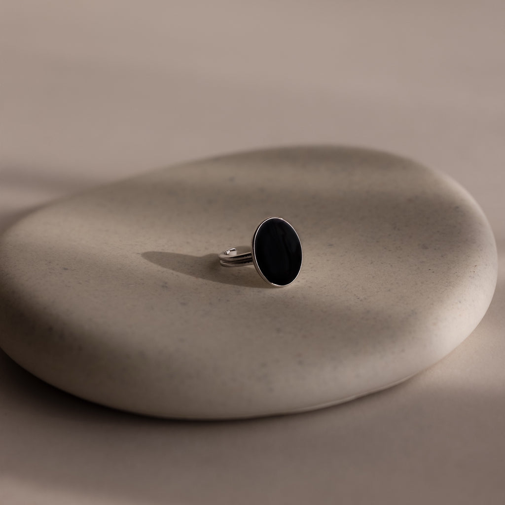 The Midnight Hour Onyx Ring, featuring a silver band and dark oval stone, is displayed on a smooth, light beige stone surface.