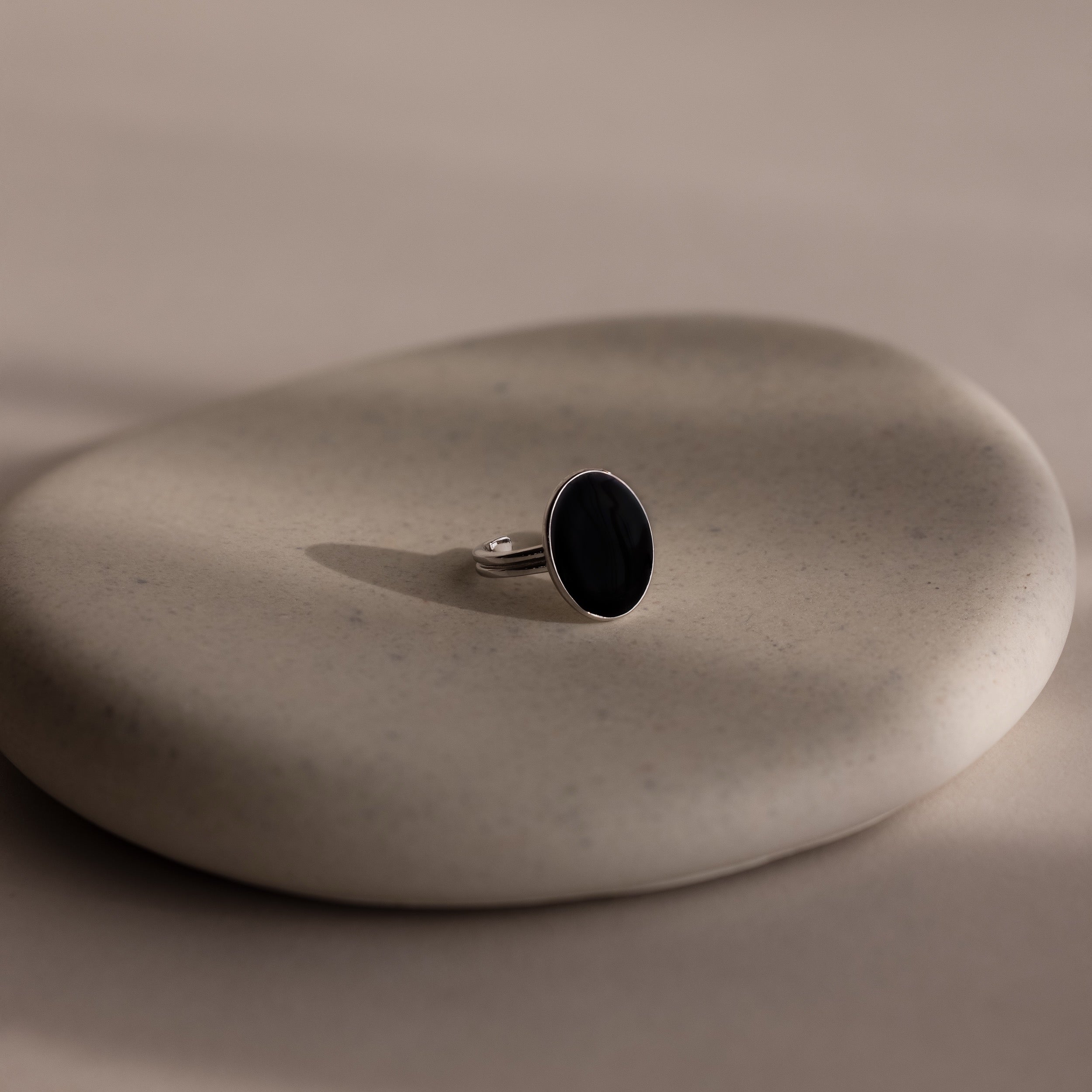 The Midnight Hour Onyx Ring, featuring a silver band and dark oval stone, is displayed on a smooth, light beige stone surface.