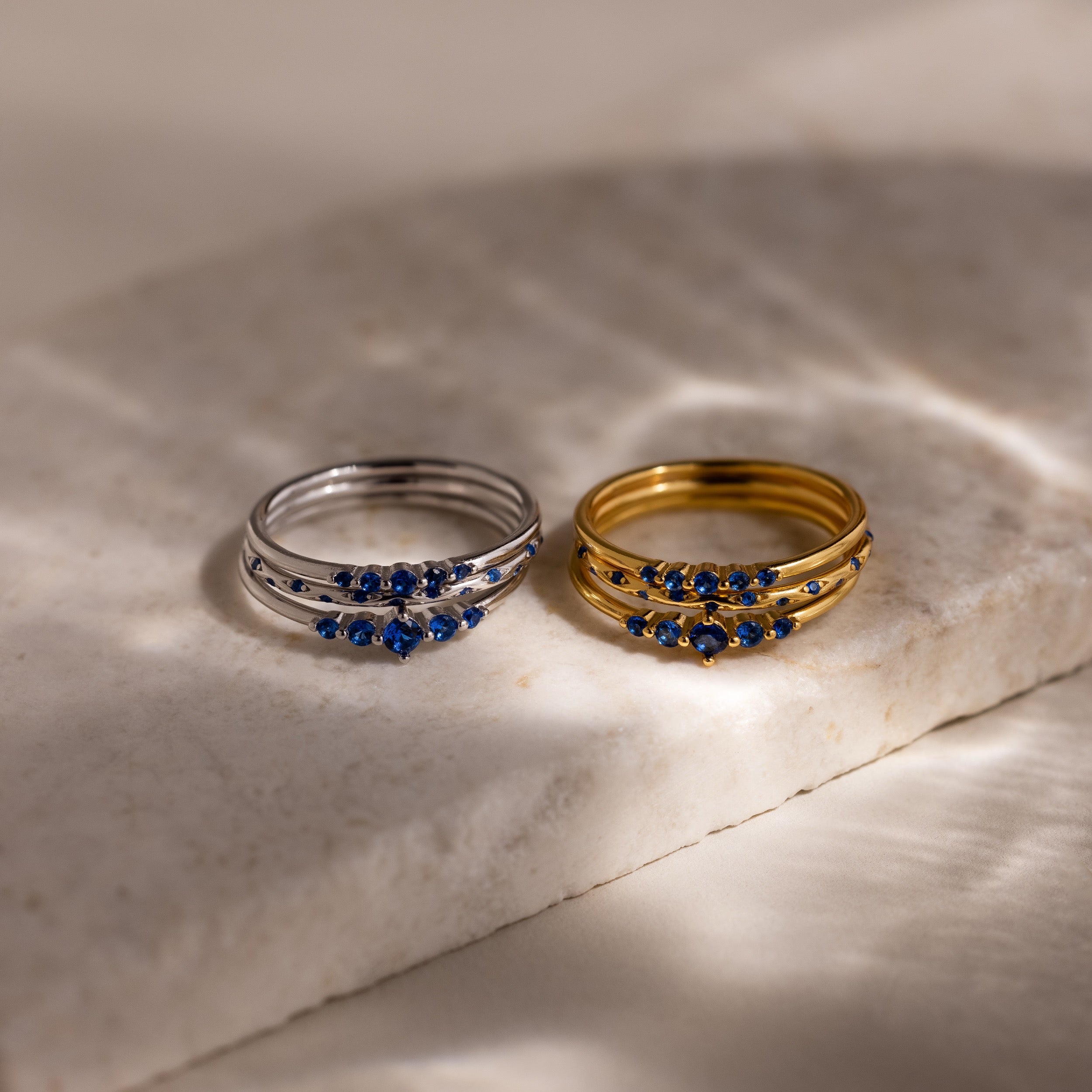 Two sets of 3 rings each, one set in silver and one set in gold, with blue stones on a marble surface.