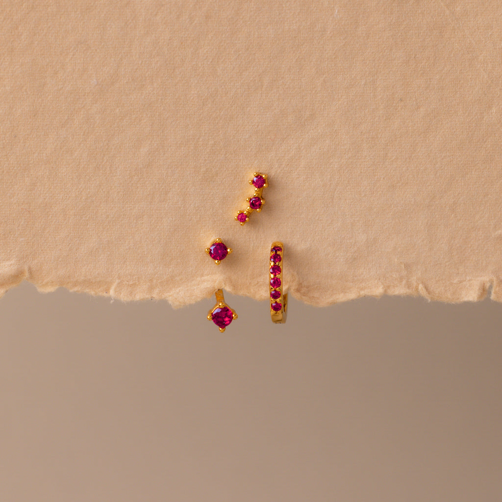 The Ruby Earring Trio Set, featuring delicate red gemstones, rests on a textured beige surface and displays its elegant charm.