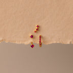 The Ruby Earring Trio Set, featuring delicate red gemstones, rests on a textured beige surface and displays its elegant charm.