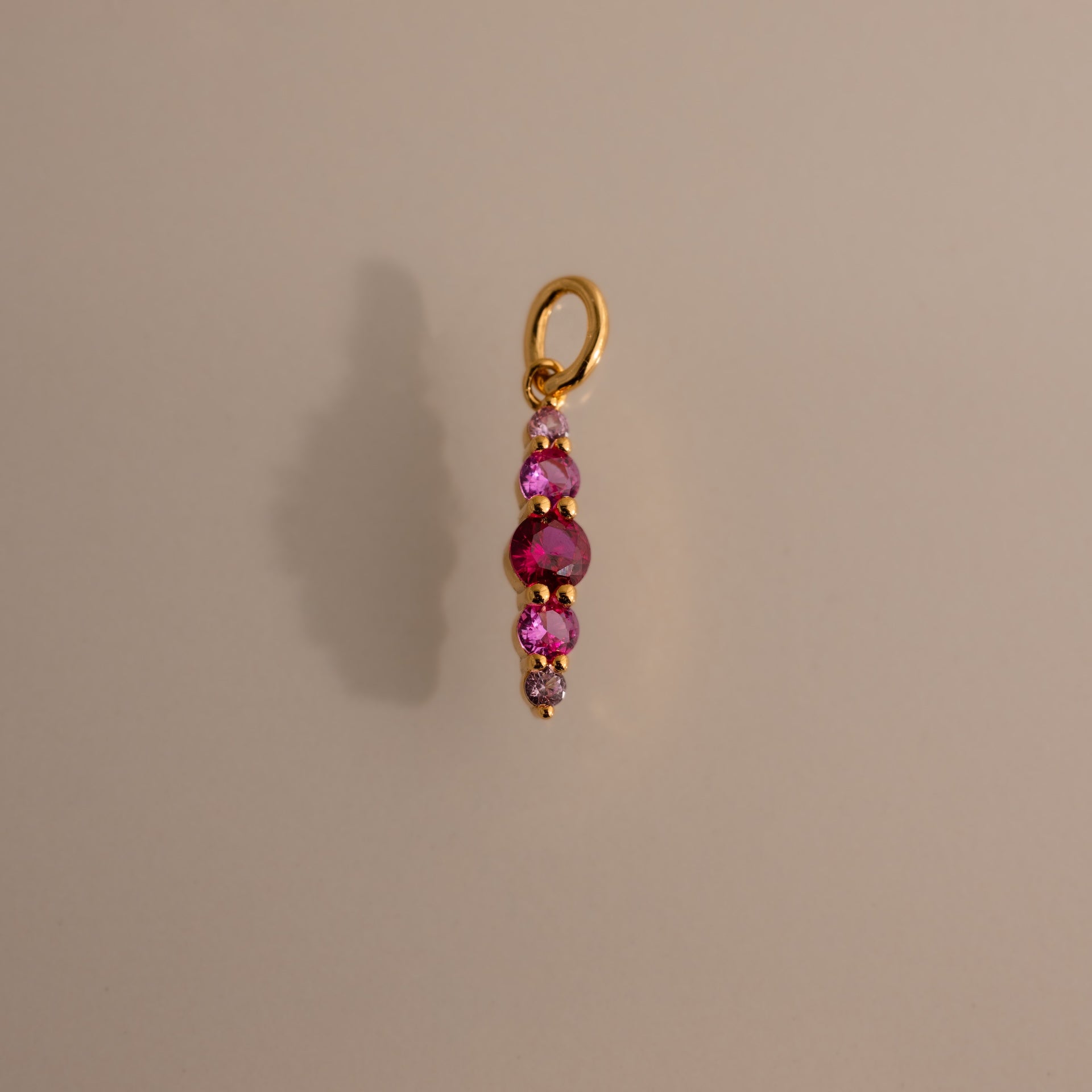 The Ombre Lace Ruby Charm is a gold pendant with four pink gemstones set vertically on a plain background.