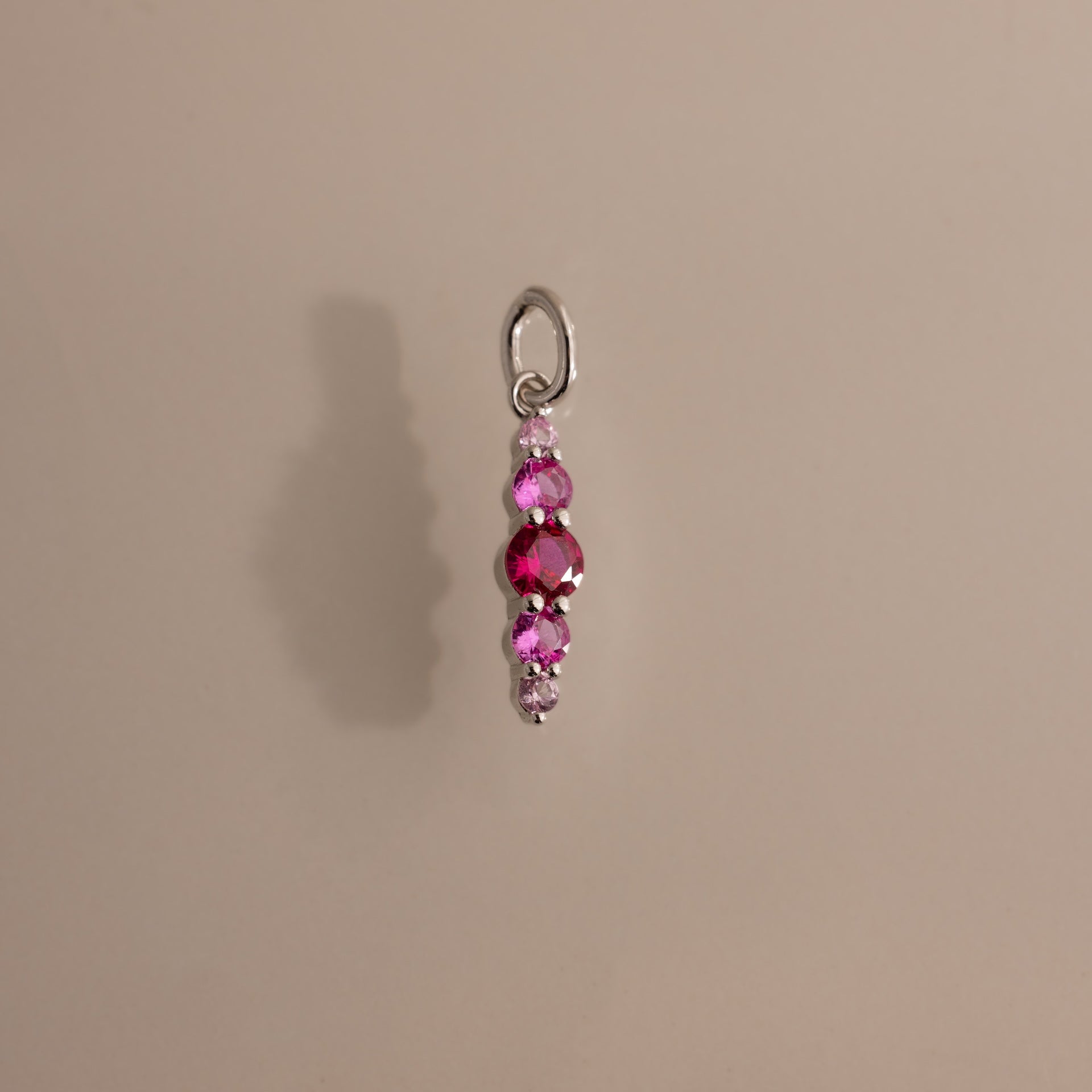 A silver pendant with three pink beads and a clear charm casts a shadow on beige—perfect as a personalized accessory or the Ombre Lace Ruby Charm.