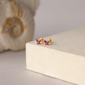 Butterfly Ruby Flatback Studs—gold butterfly-shaped earrings with pink gemstones, perfect as birthstone studs, shown on a beige surface with shell decor in the background.