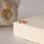 Butterfly Ruby Flatback Studs—gold butterfly-shaped earrings with pink gemstones, perfect as birthstone studs, shown on a beige surface with shell decor in the background.
