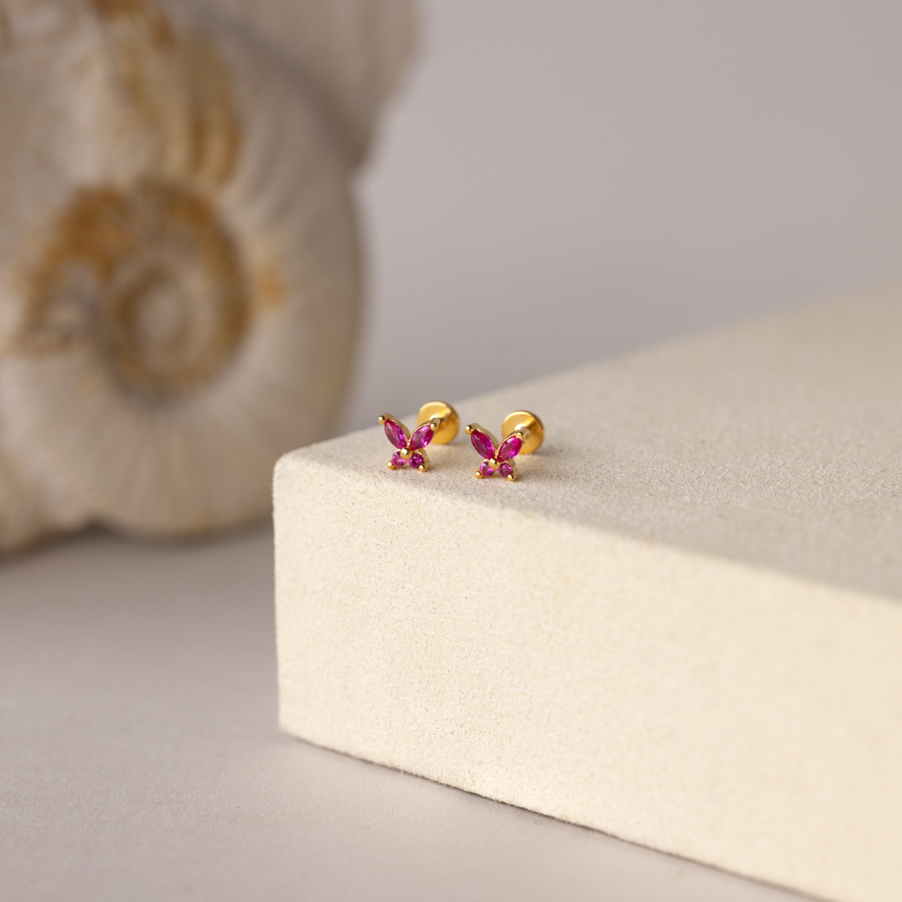 Butterfly Ruby Flatback Studs—gold butterfly-shaped earrings with pink gemstones, perfect as birthstone studs, shown on a beige surface with shell decor in the background.