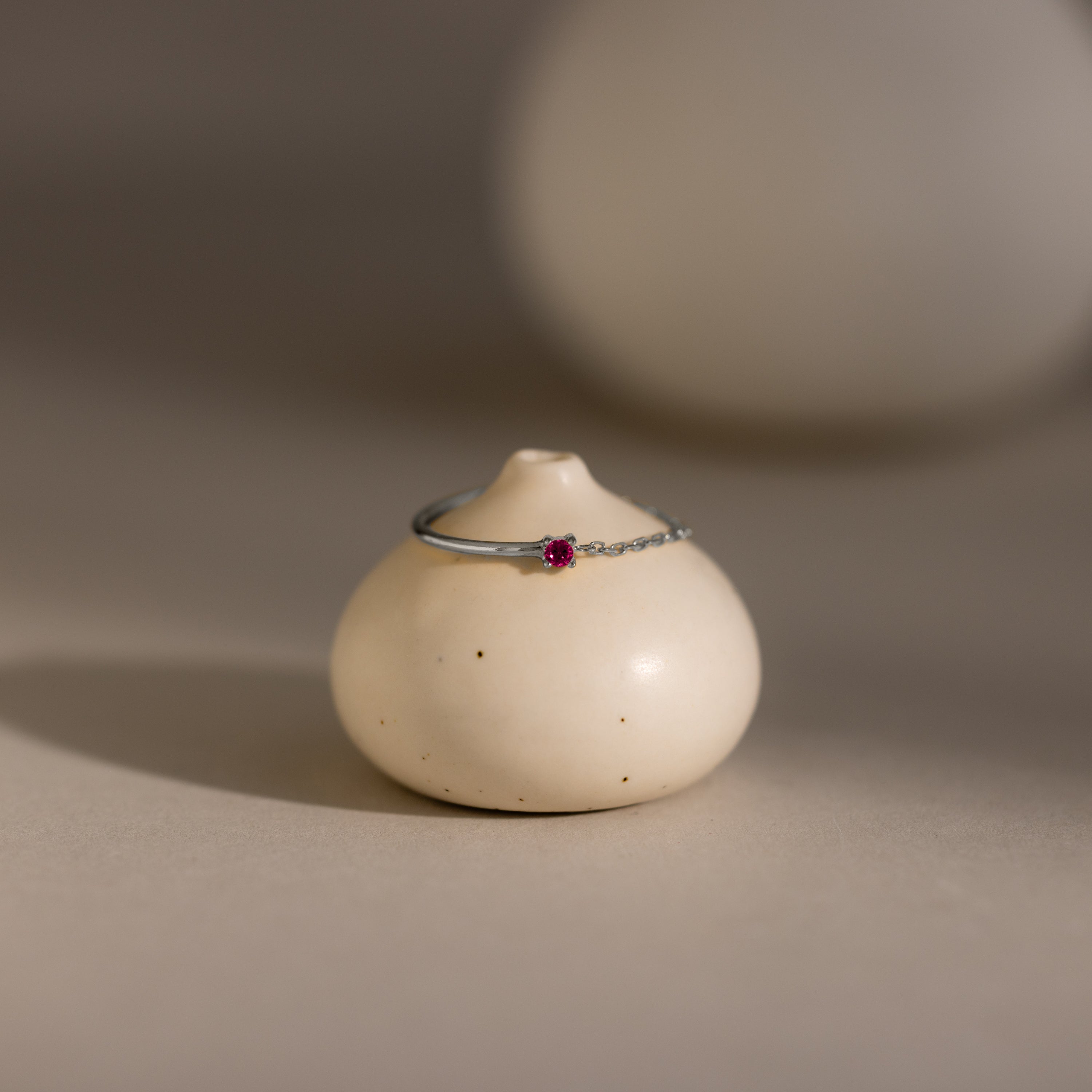 The Ruby Half Chain Ring is a delicate silver ring with a small red gemstone, ideal for dainty jewelry lovers, shown on a round cream ceramic stand.