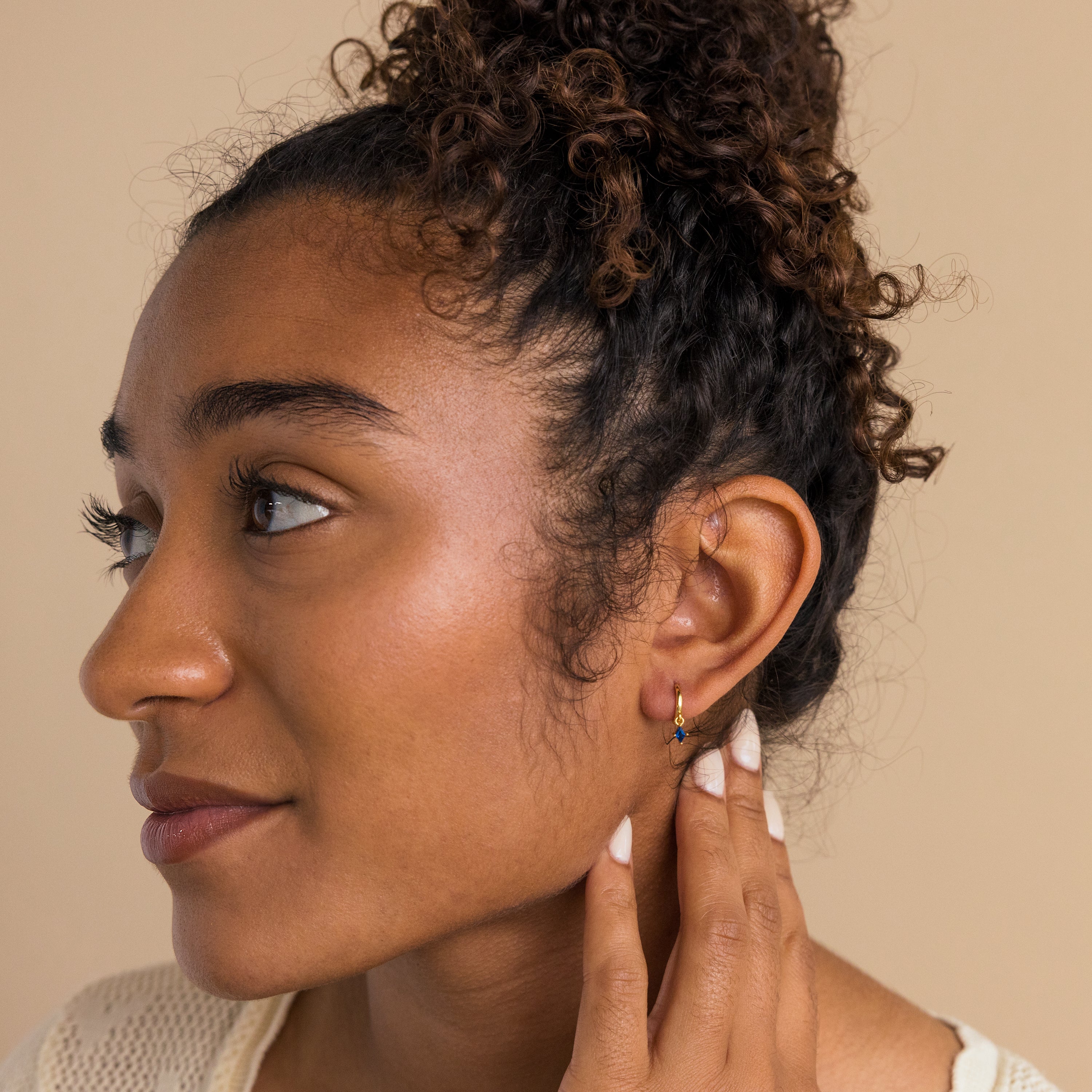Woman with curly hair in a bun touches her ear, showing off the Sapphire Drop Huggies while looking to the left.