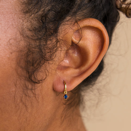 Close-up of an ear wearing Birthstone Drop Huggies—small gold hoop earrings with a blue gemstone pendant—for a touch of personalized, elegant jewelry.