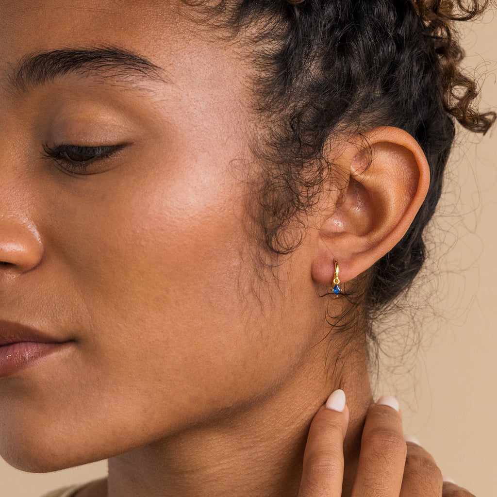 Close-up of a woman wearing dainty earrings—a small gold hoop with a red ruby pendant, eyes closed and hand near her face. Discover our Ruby Drop Huggies for personalized jewelry style.