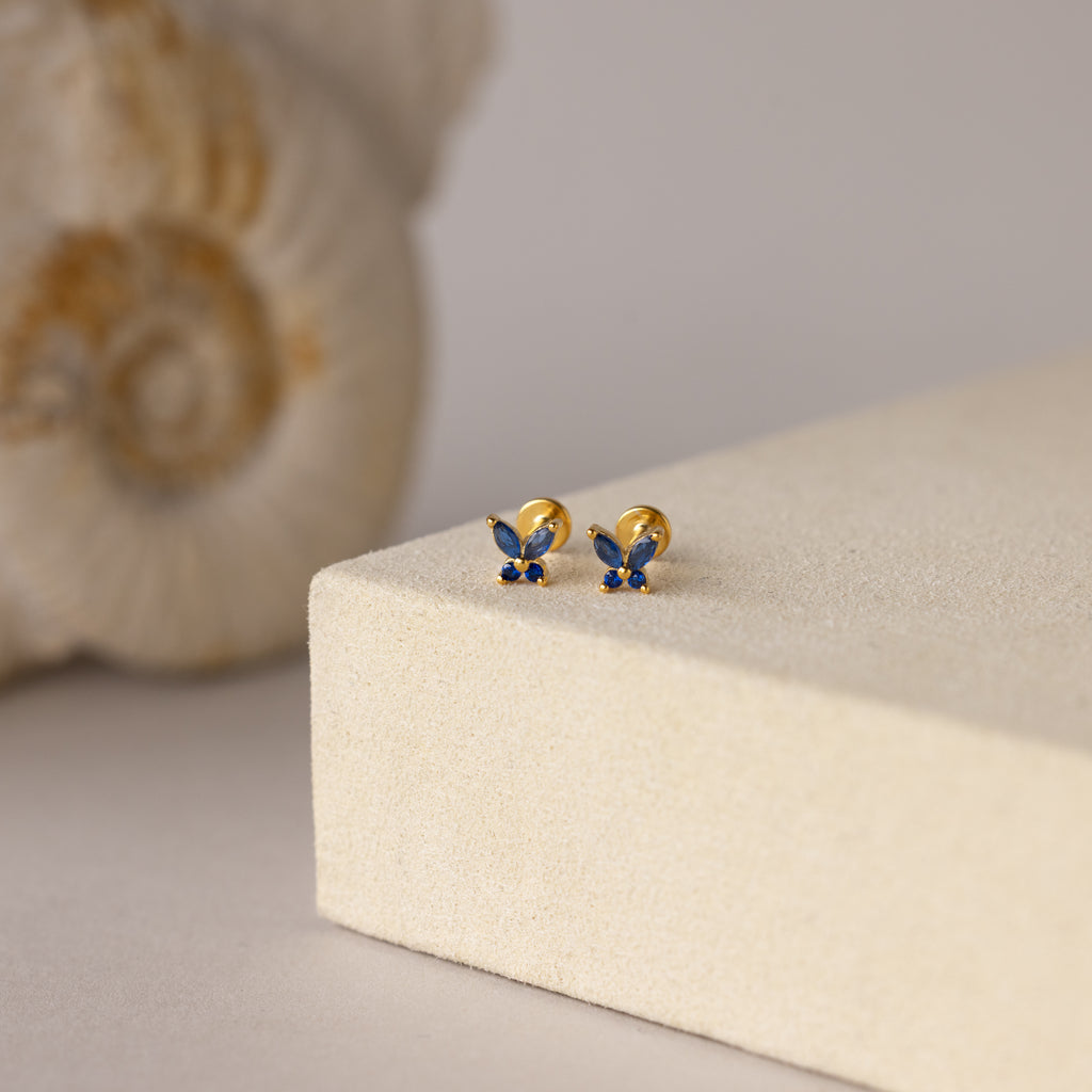 Butterfly Sapphire Flatback Studs with blue gemstones rest on a beige block, seashell in the background—a charming personalized gift.