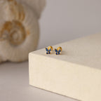 Butterfly Sapphire Flatback Studs with blue gemstones rest on a beige block, seashell in the background—a charming personalized gift.