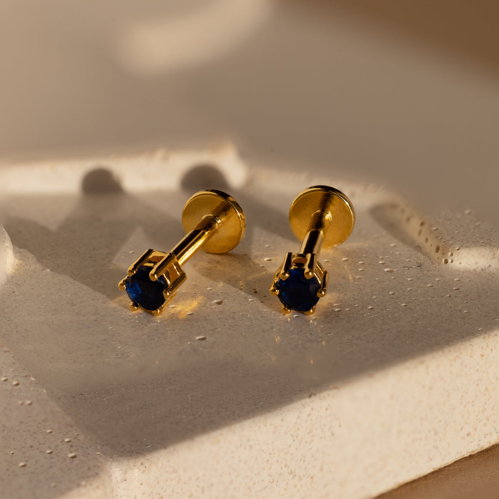 Sapphire Flatback Studs with deep blue stones sit on a textured, cream-colored surface in soft light, making them perfect as birthstone earrings.