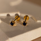 Sapphire Flatback Studs with deep blue stones sit on a textured, cream-colored surface in soft light, making them perfect as birthstone earrings.