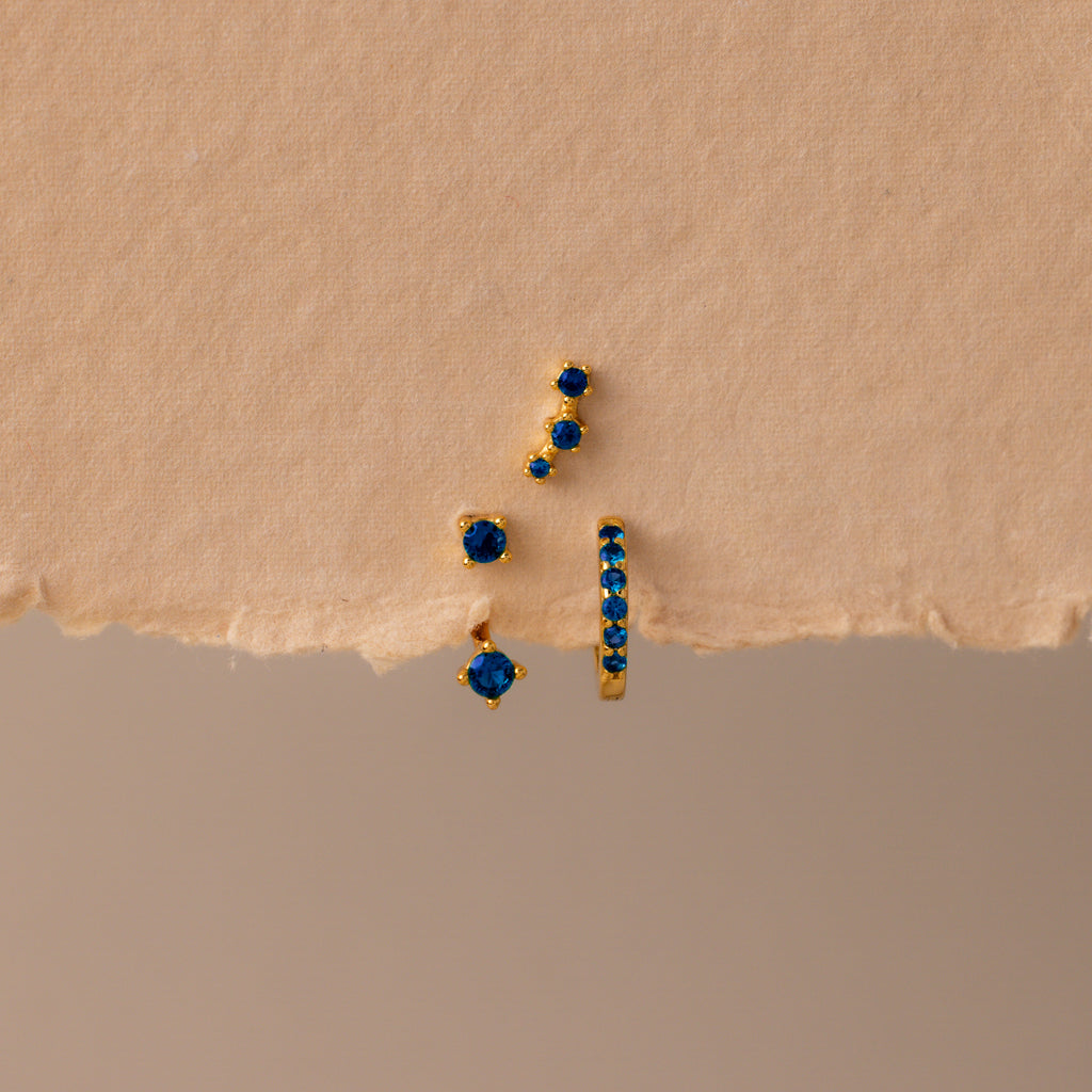The Sapphire Earring Trio Set features four gold birthstone earrings with small blue gemstones, including a pavé huggie hoop, displayed on a textured beige surface.