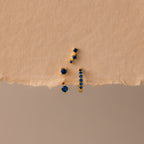 The Sapphire Earring Trio Set features four gold birthstone earrings with small blue gemstones, including a pavé huggie hoop, displayed on a textured beige surface.