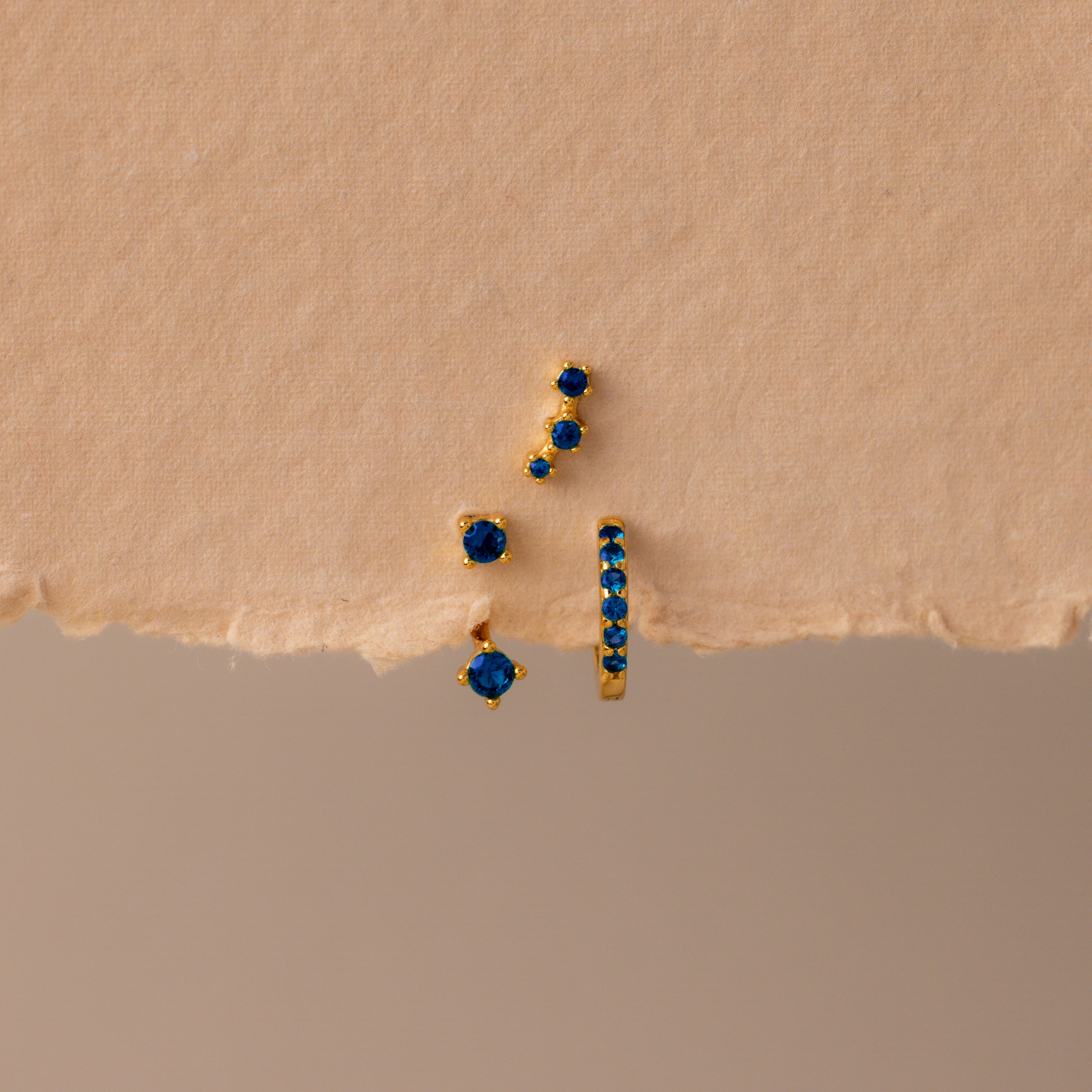 The Sapphire Earring Trio Set features four gold birthstone earrings with small blue gemstones, including a pavé huggie hoop, displayed on a textured beige surface.