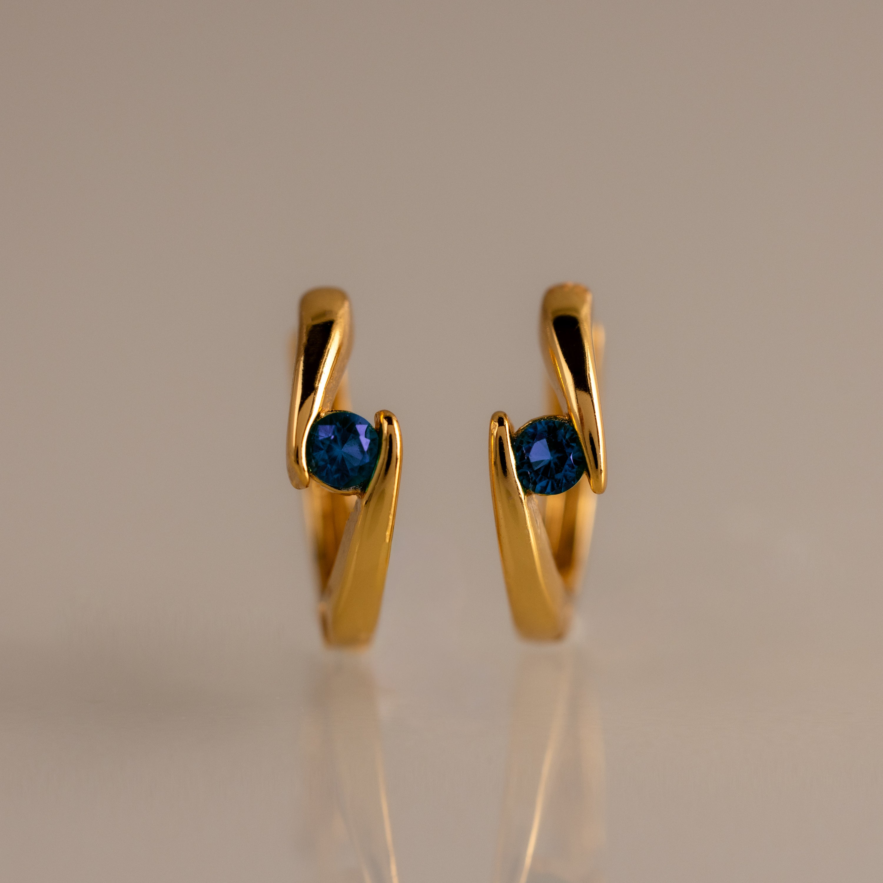 Floating Sapphire Huggies—gold hoop earrings with blue gemstones—stand upright on a neutral, reflective surface, making them the perfect personalized jewelry piece.
