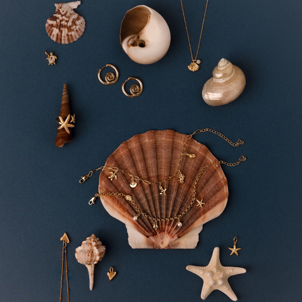 The Seaside Pearl Charm Bracelet in 18K Gold displayed on seashells and a starfish against a dark blue background evokes ocean-inspired, beachy jewelry vibes.