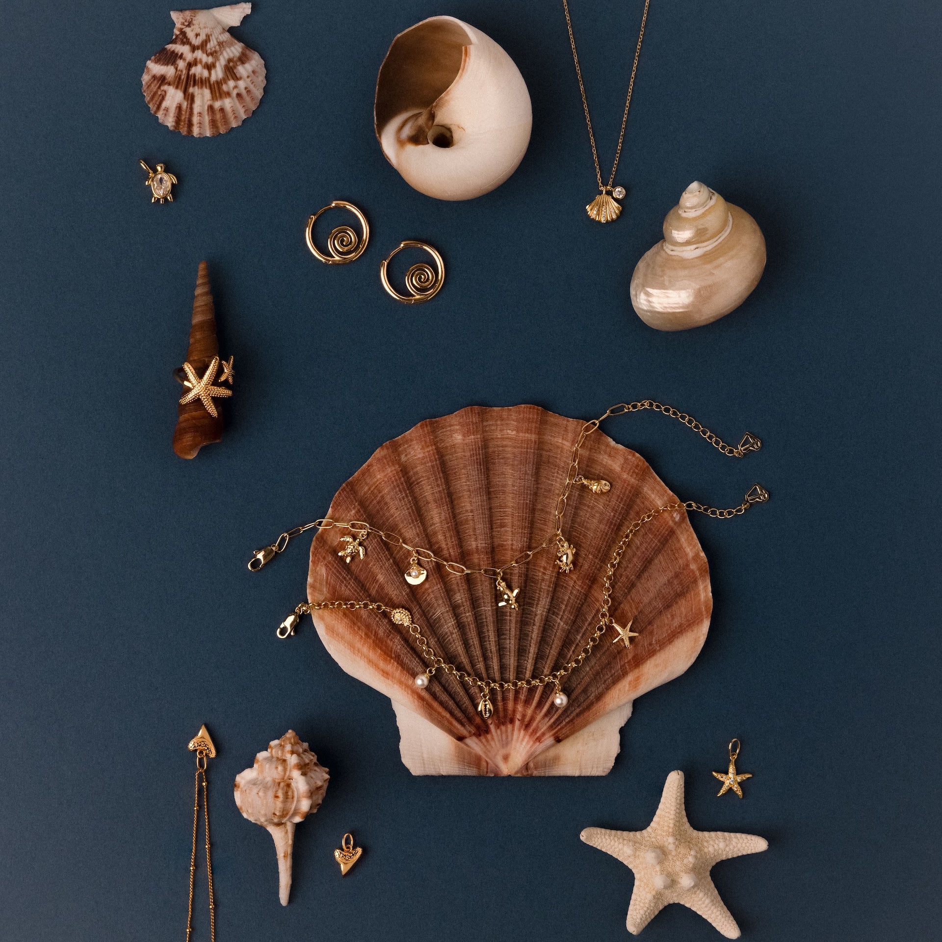 The Seaside Pearl Charm Bracelet in 18K Gold displayed on seashells and a starfish against a dark blue background evokes ocean-inspired, beachy jewelry vibes.