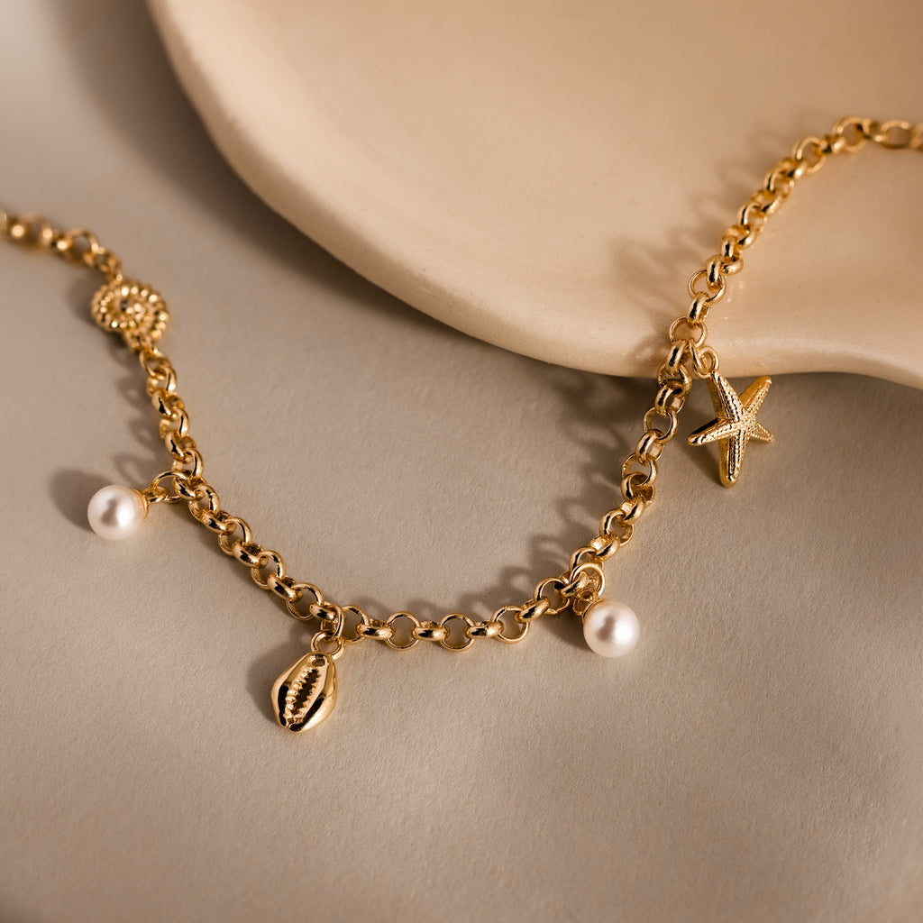 The Seaside Pearl Charm Bracelet in 18K Gold features ocean-inspired pearl, starfish, shell, and bead charms for elegant beachy style on a beige surface.