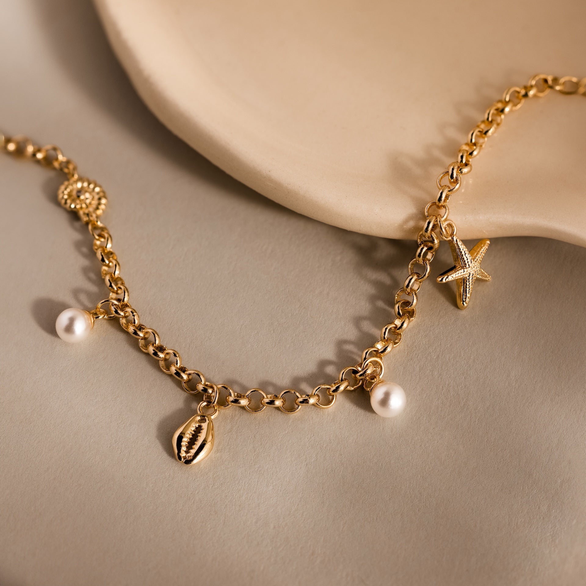 The Seaside Pearl Charm Bracelet in 18K Gold features ocean-inspired pearl, starfish, shell, and bead charms for elegant beachy style on a beige surface.