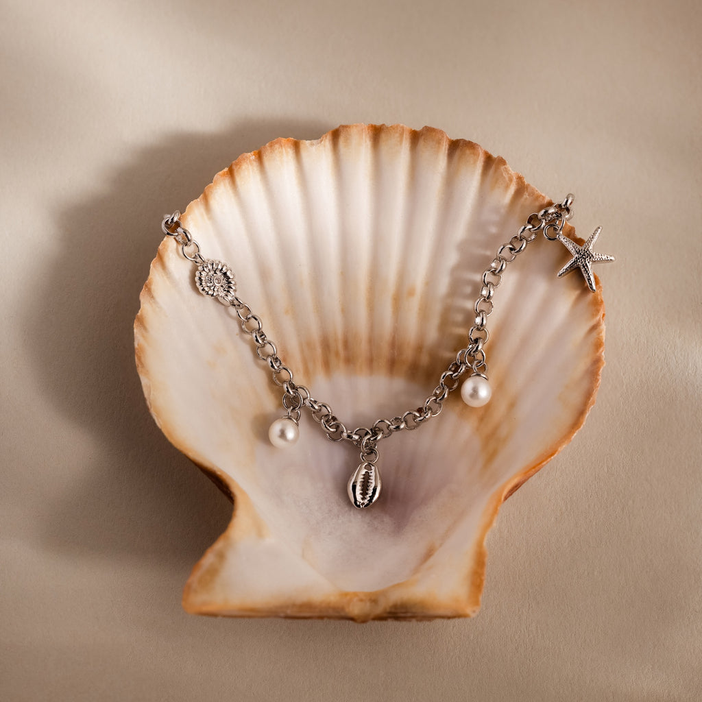 The Seaside Pearl Charm Bracelet in Silver, adorned with ocean-themed charms, is displayed in an open seashell on a beige surface.