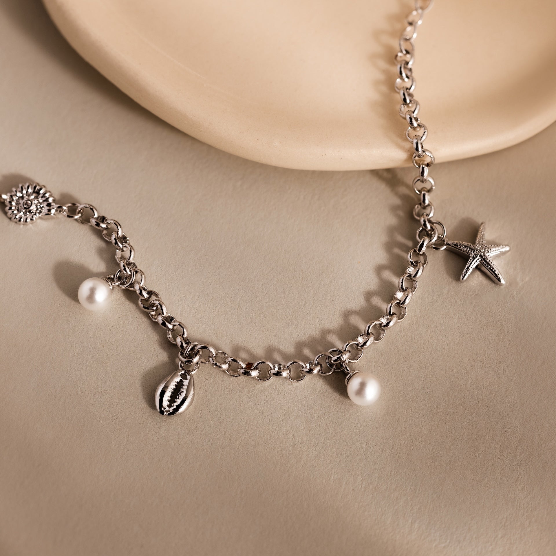 The Seaside Pearl Charm Bracelet with pearl, starfish, and shell charms is displayed on a beige surface beside a round dish—an ideal piece of coastal jewelry.