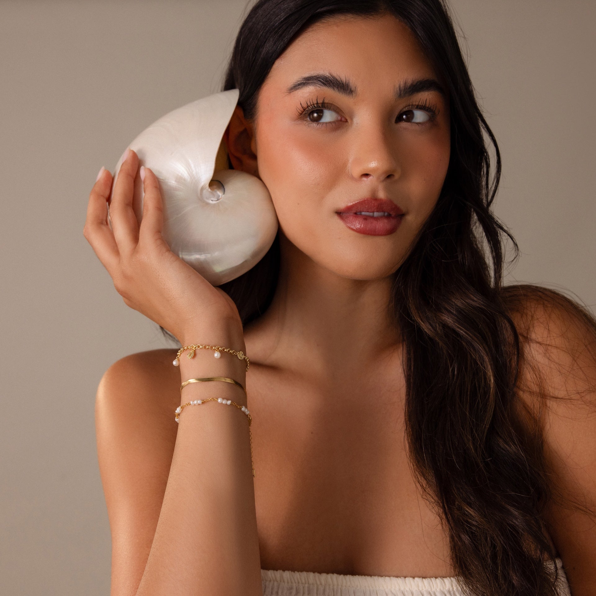 A woman with long dark hair holds a seashell to her ear, wearing beachy jewelry and the Seaside Pearl Charm Bracelet in 18K Gold, adorned with ocean-inspired charms.