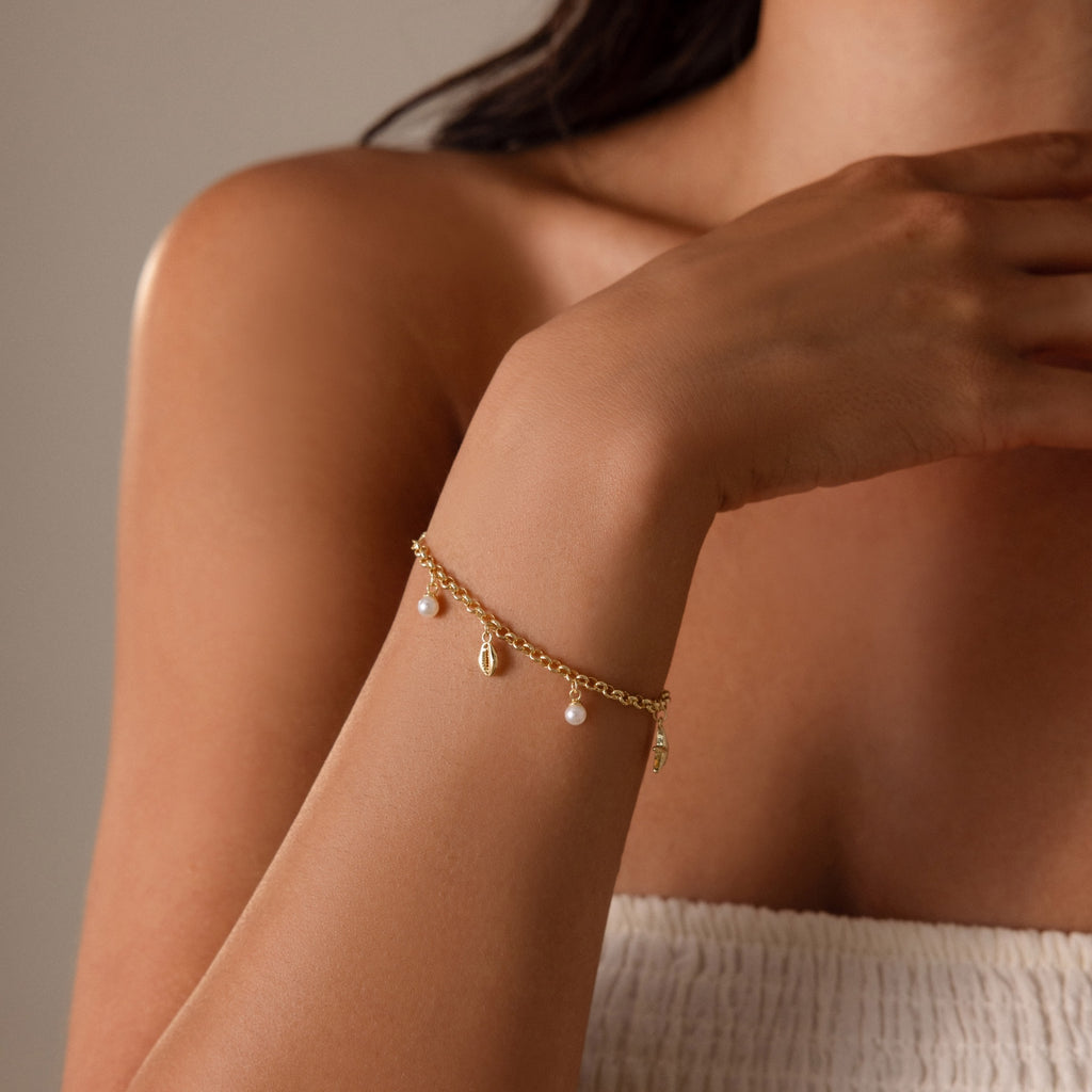 A woman in a white top wears the Seaside Pearl Charm Bracelet in 18K Gold, featuring ocean-inspired charms and small pearls on her wrist.