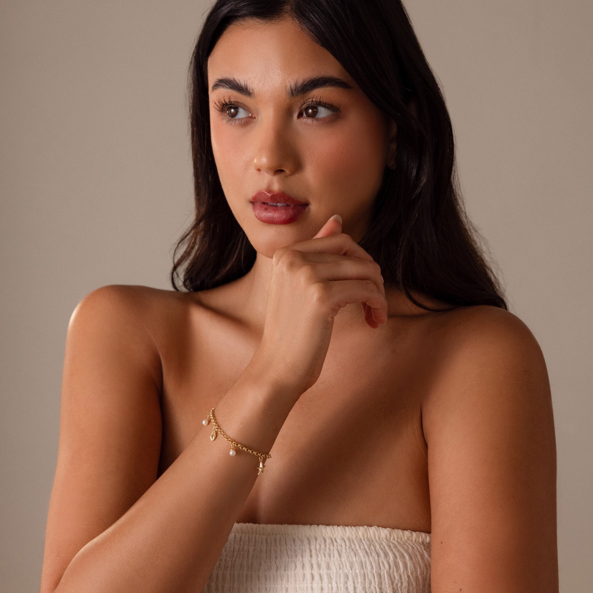 Woman with long dark hair wearing a strapless top and the Seaside Pearl Charm Bracelet in 18K Gold, gazing thoughtfully to the side and embodying beach-inspired elegance.