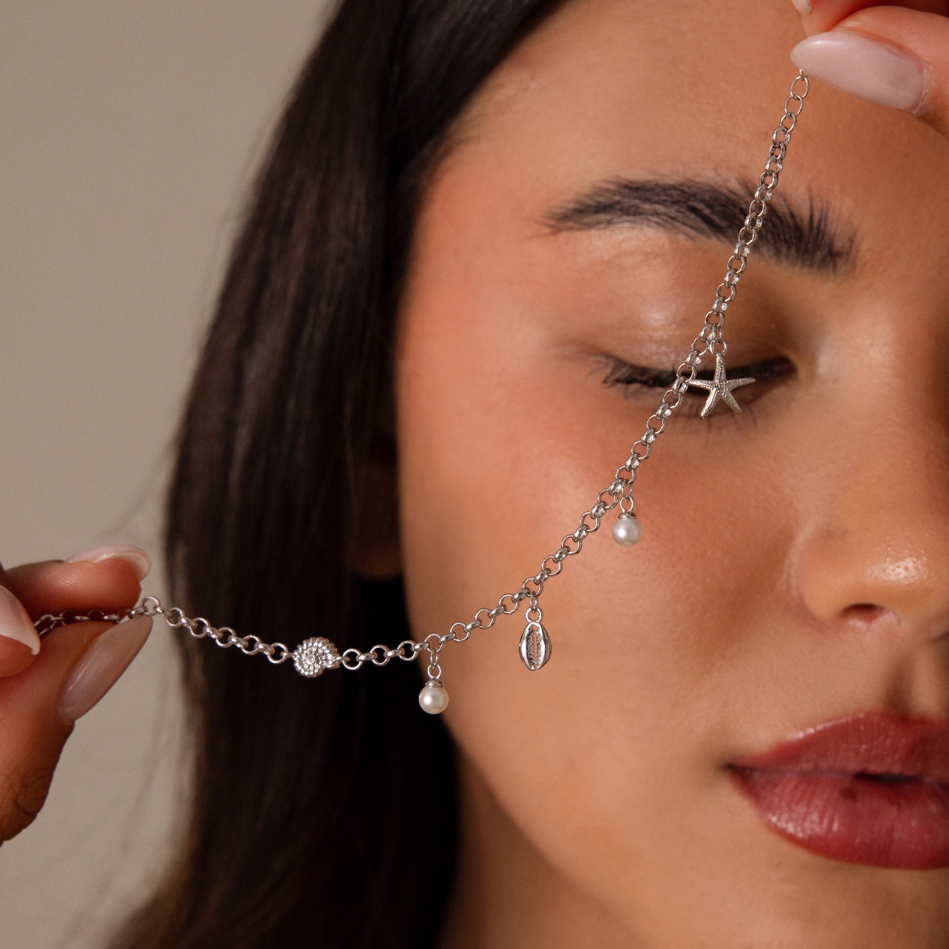 A woman holds the Seaside Pearl Charm Bracelet, featuring ocean-inspired charms, pearls, and starfish details, near her face with her eyes closed.