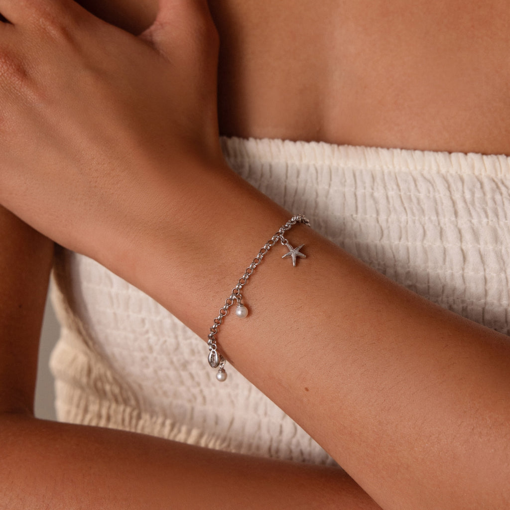 The Seaside Pearl Charm Bracelet in Silver, adorned with ocean-themed charms like starfish and pearls, stands out on a woman’s wrist as she wears a white textured top.