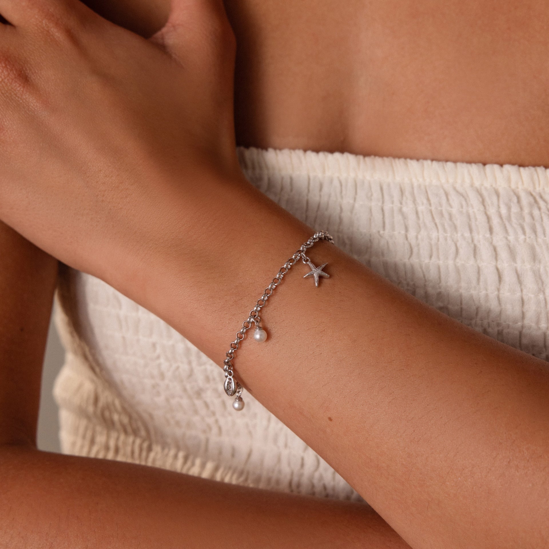 The Seaside Pearl Charm Bracelet in Silver, adorned with ocean-themed charms like starfish and pearls, stands out on a woman’s wrist as she wears a white textured top.