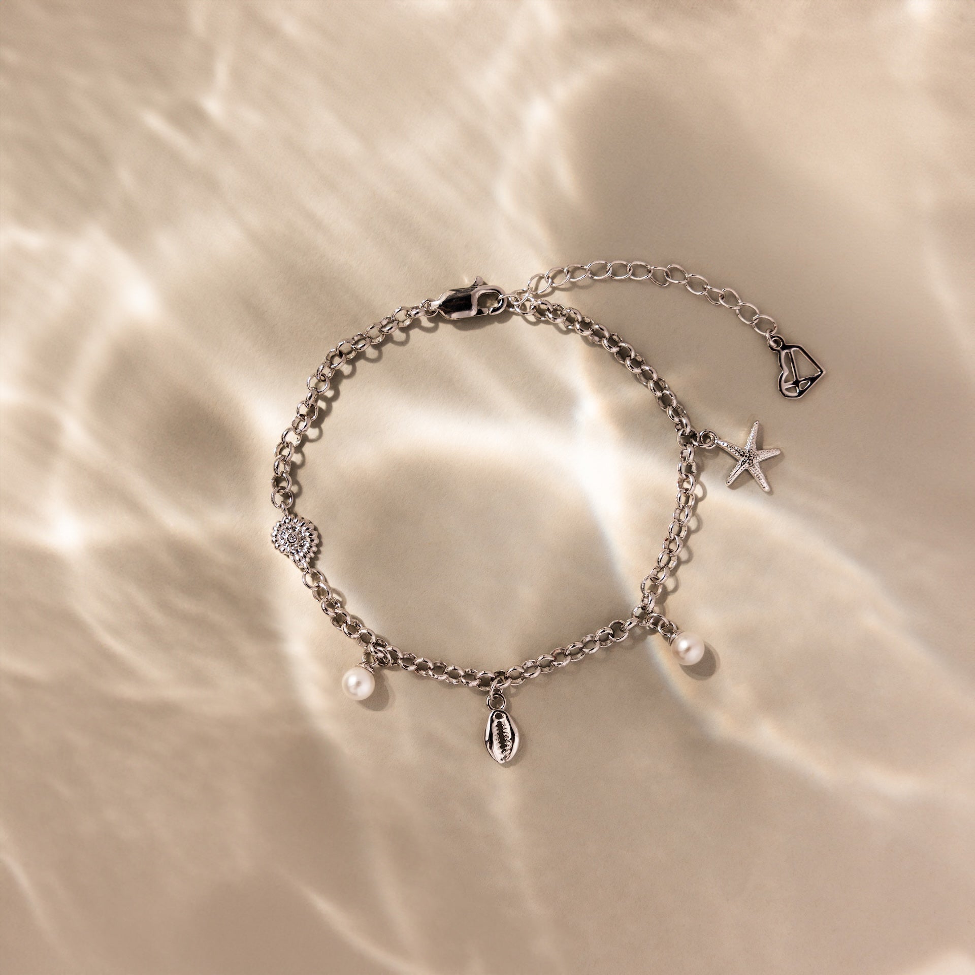 The Seaside Pearl Charm Bracelet in Silver, adorned with ocean-inspired pearl, shell, and starfish charms, gleams on a sunny beach—perfect for coastal jewelry lovers.