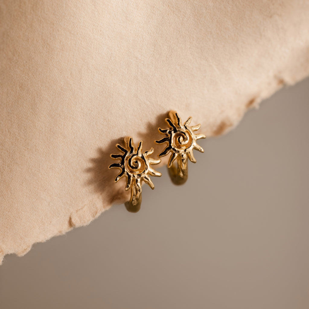 Celestial Sun Huggies in 18K Gold shine against a textured beige surface and soft, neutral background, making these sun-shaped earrings perfect statement jewelry for any occasion.
