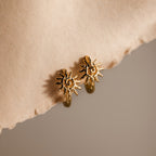 Celestial Sun Huggies in 18K Gold shine against a textured beige surface and soft, neutral background, making these sun-shaped earrings perfect statement jewelry for any occasion.