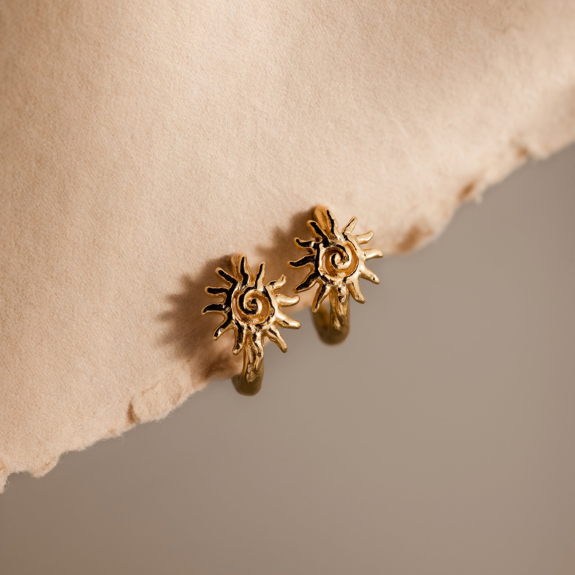 Celestial Sun Huggies in 18K Gold shine against a textured beige surface and soft, neutral background, making these sun-shaped earrings perfect statement jewelry for any occasion.