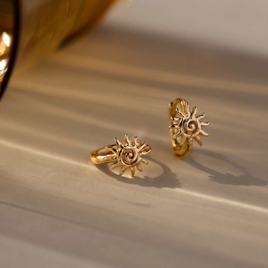 Two Celestial Sun Huggies in 18K Gold with sun-shaped designs sit on a beige surface under warm lighting—ideal for anyone who loves striking statement jewelry.