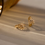 Two Celestial Sun Huggies in 18K Gold with sun-shaped designs sit on a beige surface under warm lighting—ideal for anyone who loves striking statement jewelry.