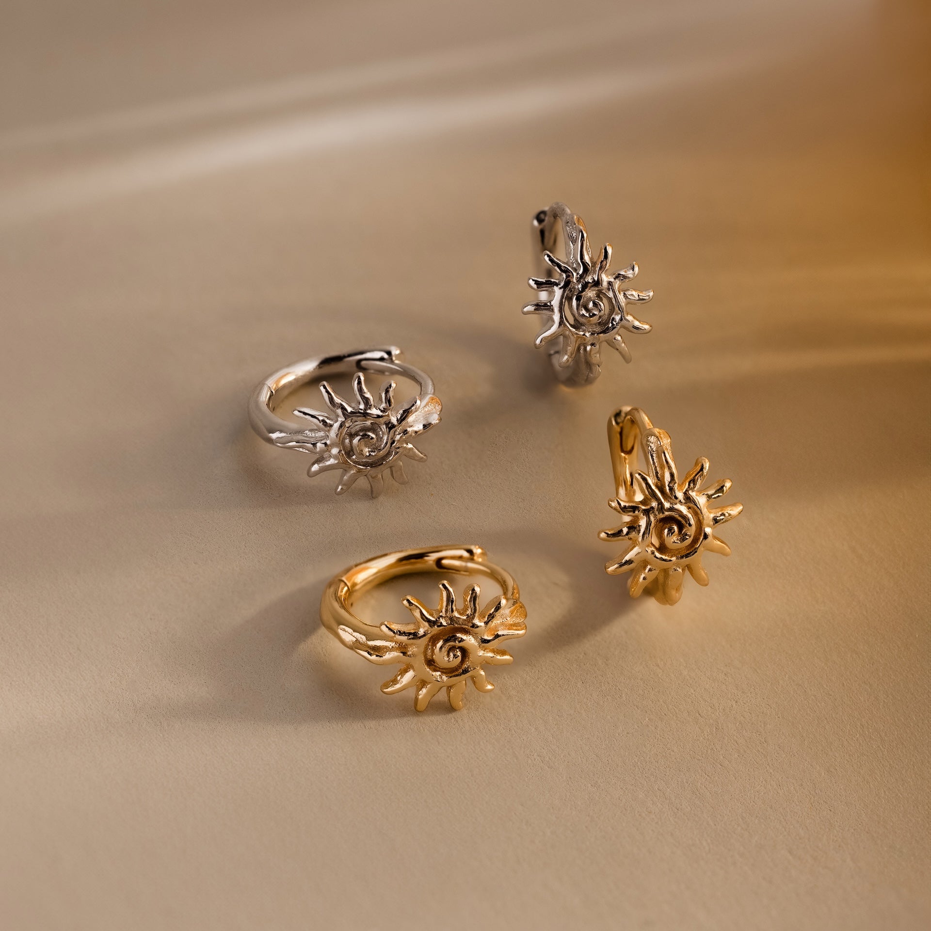 Two pairs of Celestial Sun Huggies—gold and silver—gleam as statement jewelry on a beige surface under warm light.