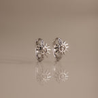 Celestial Sun Huggies in Silver feature sunburst designs and upright silver hoops, reflected on a smooth beige surface—stunning statement jewelry to elevate any look.