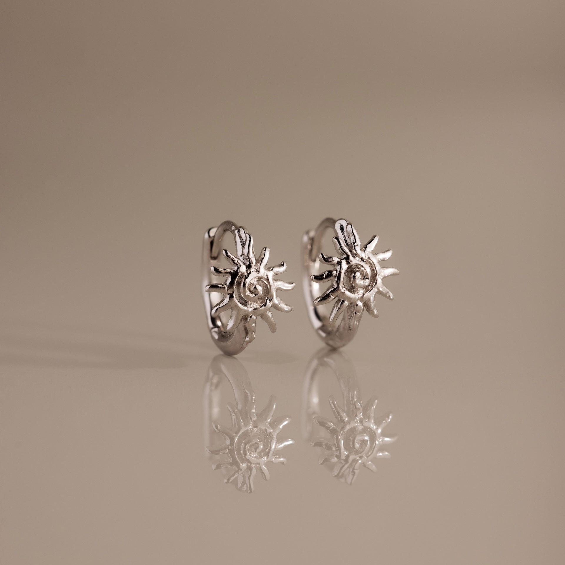 Celestial Sun Huggies in Silver feature sunburst designs and upright silver hoops, reflected on a smooth beige surface—stunning statement jewelry to elevate any look.