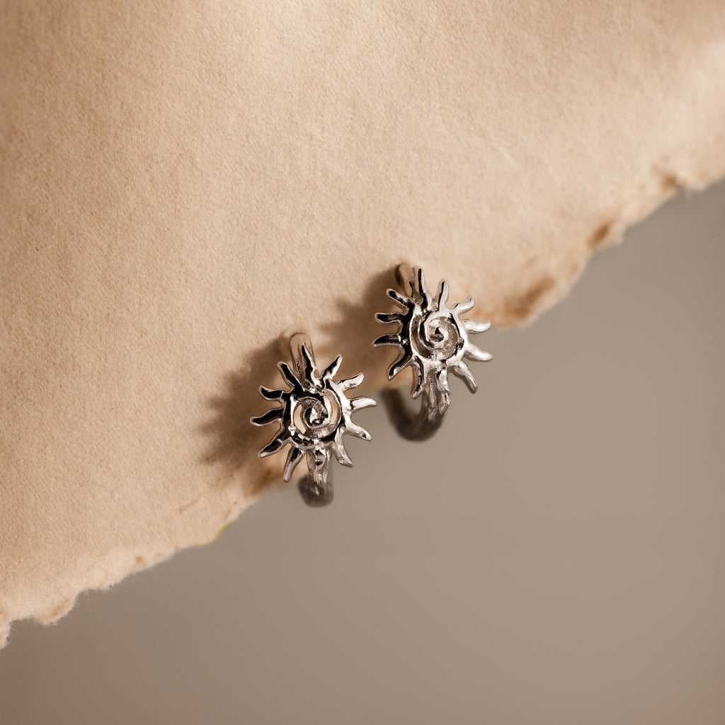Celestial Sun Huggies in Silver, featuring a radiant sunburst design, are displayed on textured beige paper—perfect statement jewelry for any occasion.
